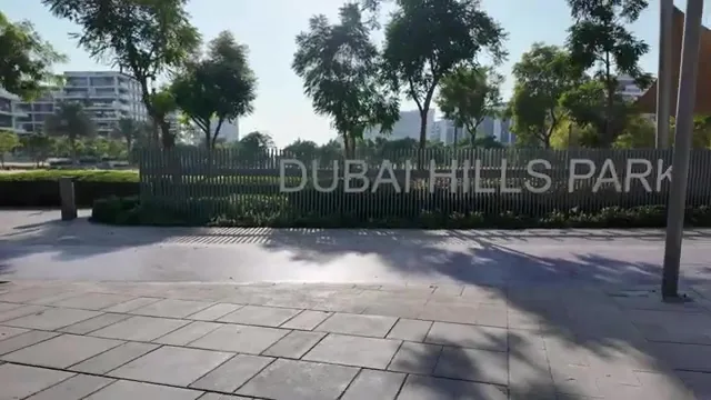 Указатель Dubai Hills Park на деревянном заборе, окруженный зелеными деревьями и кустарниками вдоль мощеной дорожки.