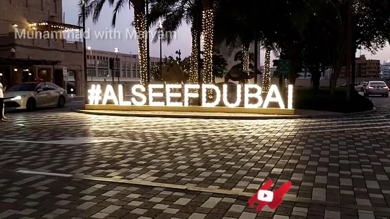 Светящаяся надпись #ALSEEFDUBAI на фоне традиционной архитектуры и подсвеченных пальм в ночное время на набережной района Аль-Сиф.