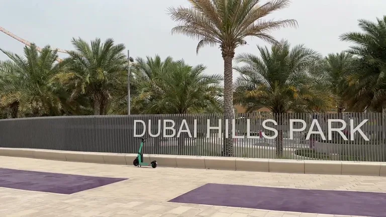 Указатель Dubai Hills Park на современном сером заборе и припаркованный рядом зеленый электросамокат.