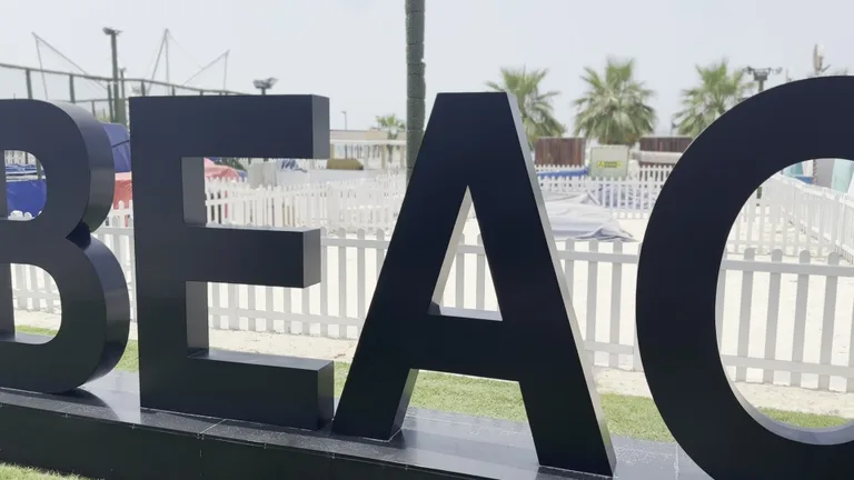 Крупный план большой черной вывески Kite Beach на фоне белого деревянного забора и пальм.